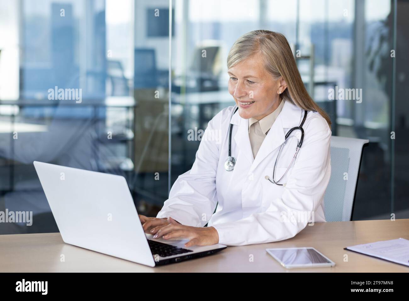 Eine ältere lächelnde grauhaarige Ärztin berät, arbeitet und schreibt online auf einem Laptop. Am Schreibtisch im Büro der Klinik sitzen. Stockfoto