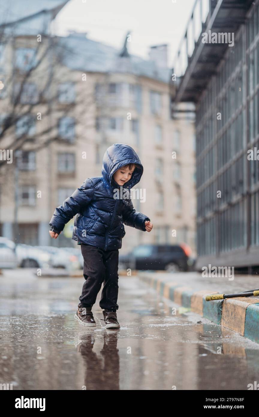 Süßer Junge, der nach Regen auf der nassen Straße spielt Stockfoto