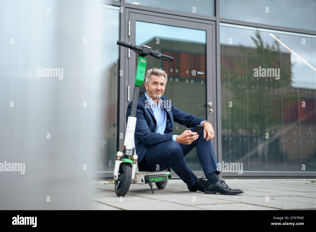 Reifer Geschäftsmann sitzt mit Smartphone auf einem elektrischen Roller Stockfoto