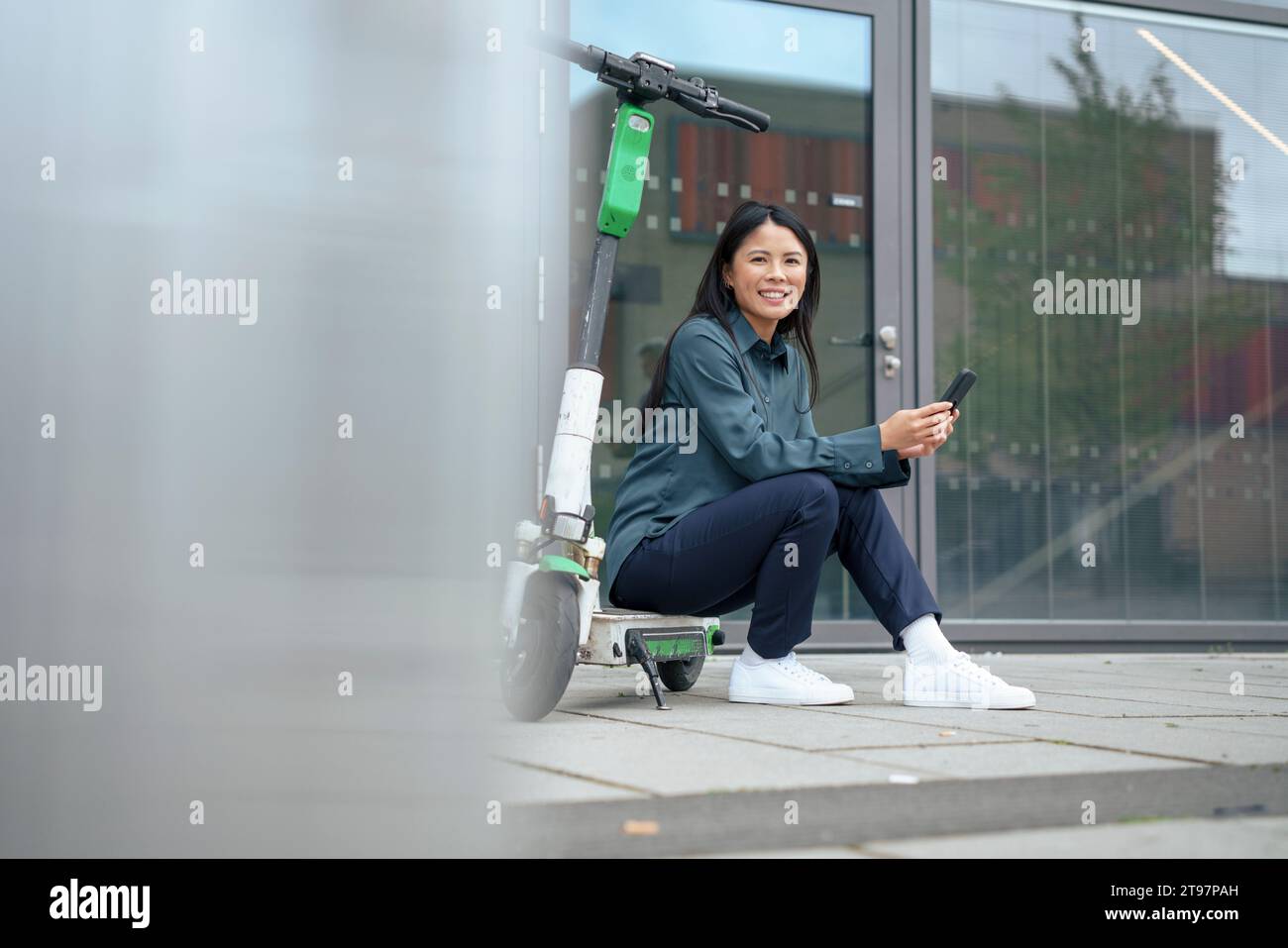Glückliche Geschäftsfrau, die auf einem elektrischen Roller sitzt, mit Smartphone auf dem Fußweg Stockfoto