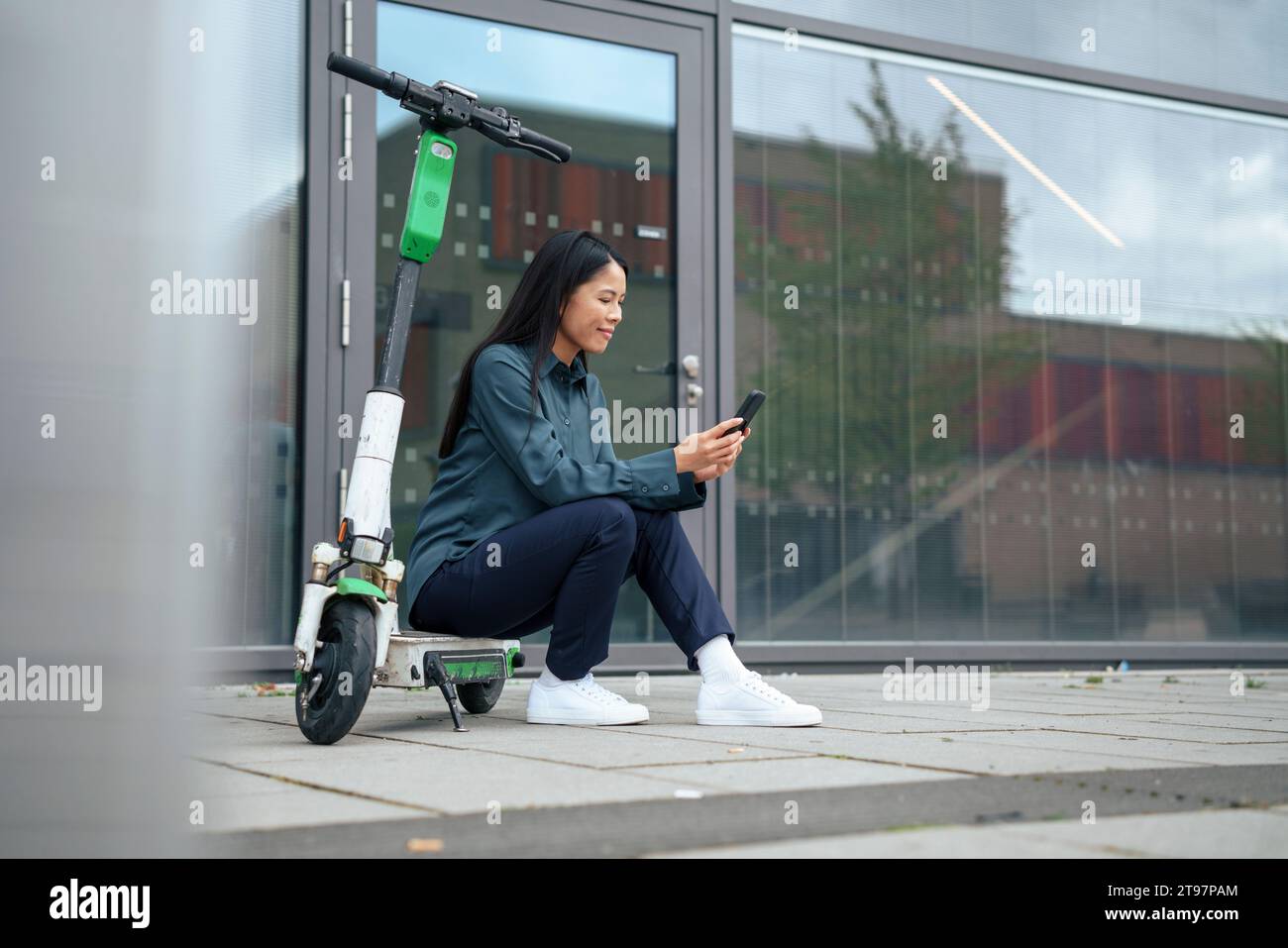 Lächelnde Geschäftsfrau, die auf einem elektrischen Roller sitzt und ein Smartphone auf dem Fußweg benutzt Stockfoto