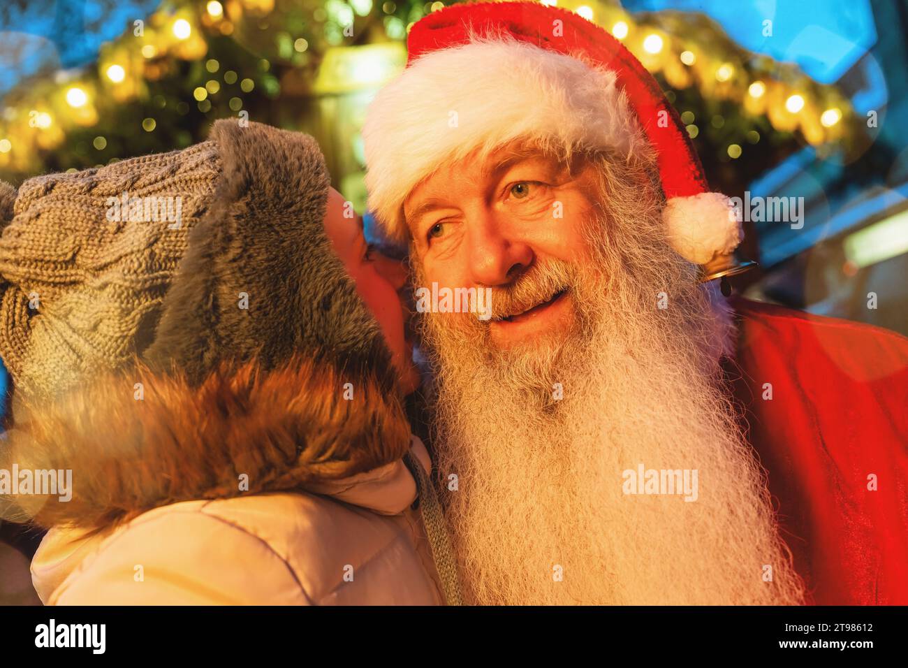 Kind flüstert dem Weihnachtsmann auf einem Weihnachtsmarkt mit festlichen Lichtern im Hintergrund Stockfoto