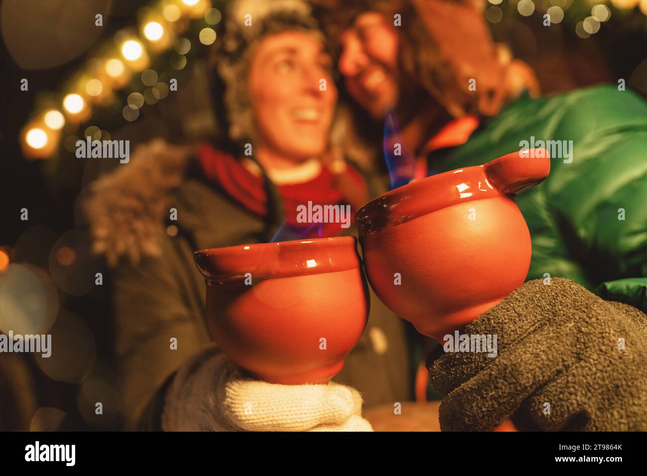 Lachende Paare genießen Feuerzangen-Punsch auf einem Weihnachtsmarkt und halten dampfende rote Tassen mit blauen Flammen. Stockfoto