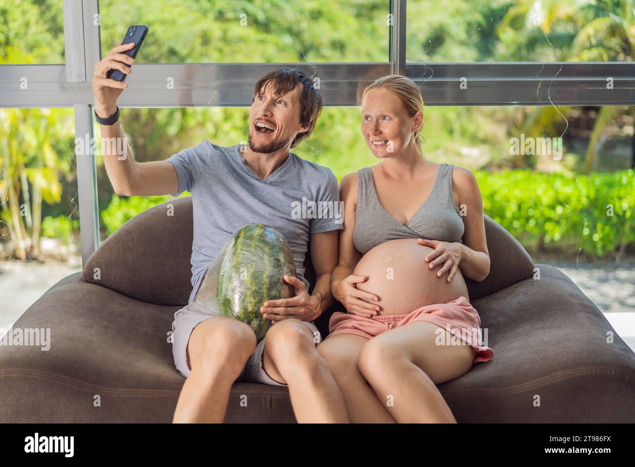 Ein humorvolles Bild: Eine schwangere Frau und ihr Ehemann benutzen spielerisch eine Wassermelone anstelle eines Bauches und zeigen komisch die Herausforderungen des Navigierens auf Stockfoto