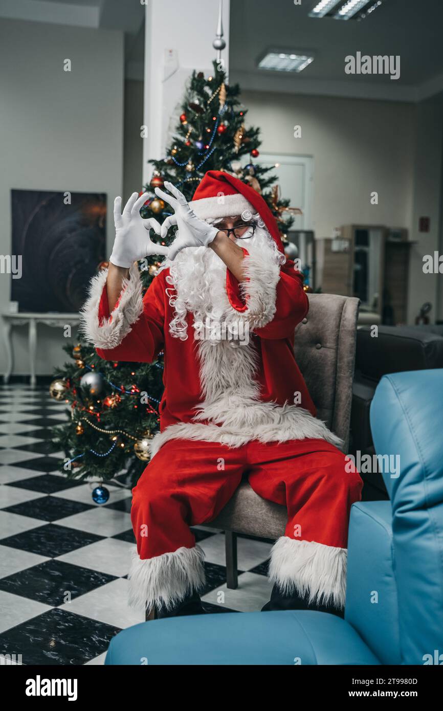 Ein fröhlicher erwachsener Mann, der ein traditionelles Weihnachtsmannskostüm trägt, sitzt vor einem geschmückten Weihnachtsbaum Stockfoto
