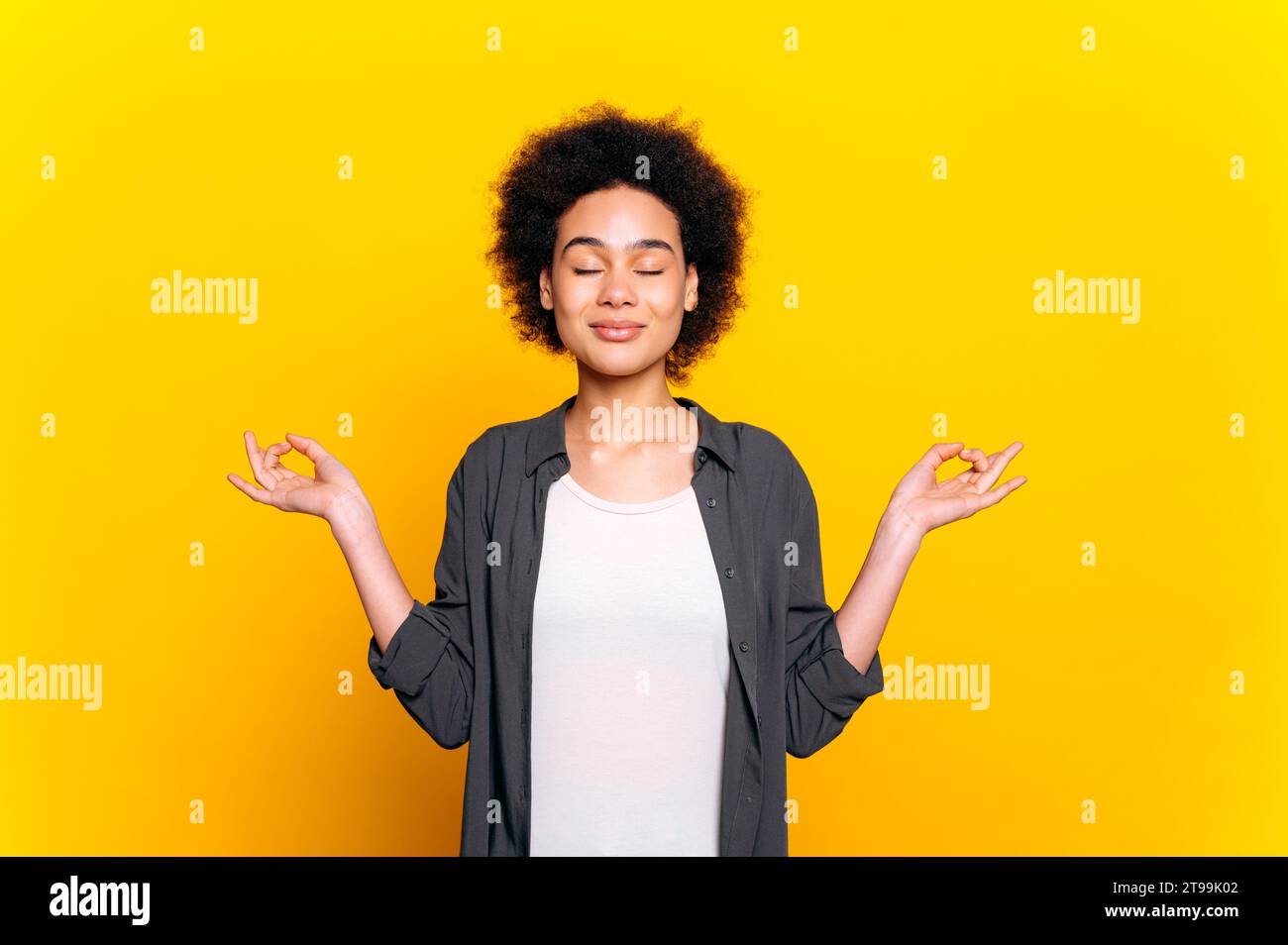 Foto einer schönen ruhigen afroamerikanischen oder brasilianischen Frau mit lockigen Haaren in einem lässigen Hemd, während sie mit geschlossenen Augen auf isoliertem gelben Hintergrund meditiert. Psychische Gesundheit, Harmonie, Entspannung Stockfoto
