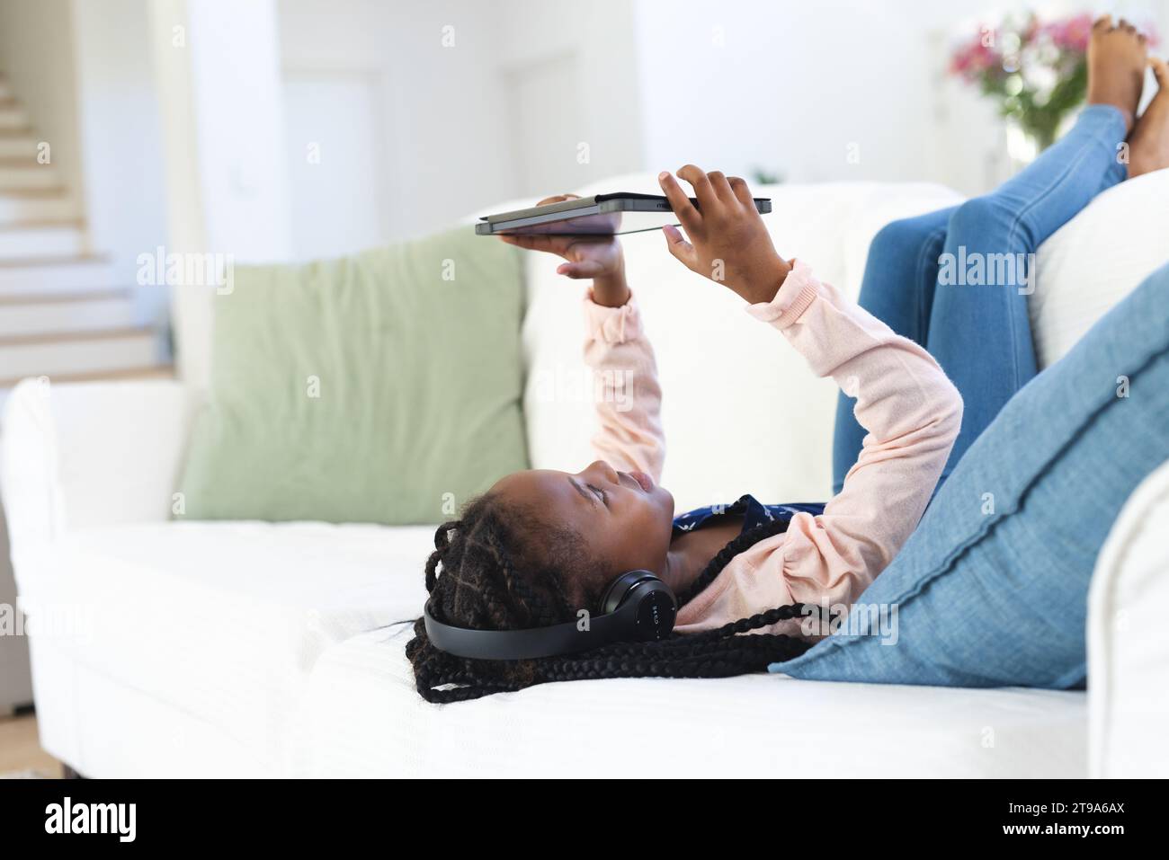 Glückliches afroamerikanisches Mädchen, das auf der Couch liegt, Musik hört und zu Hause ein Tablet benutzt, Kopierraum. Kindheit, Ausdruck, Technologie, Kommunikation Stockfoto