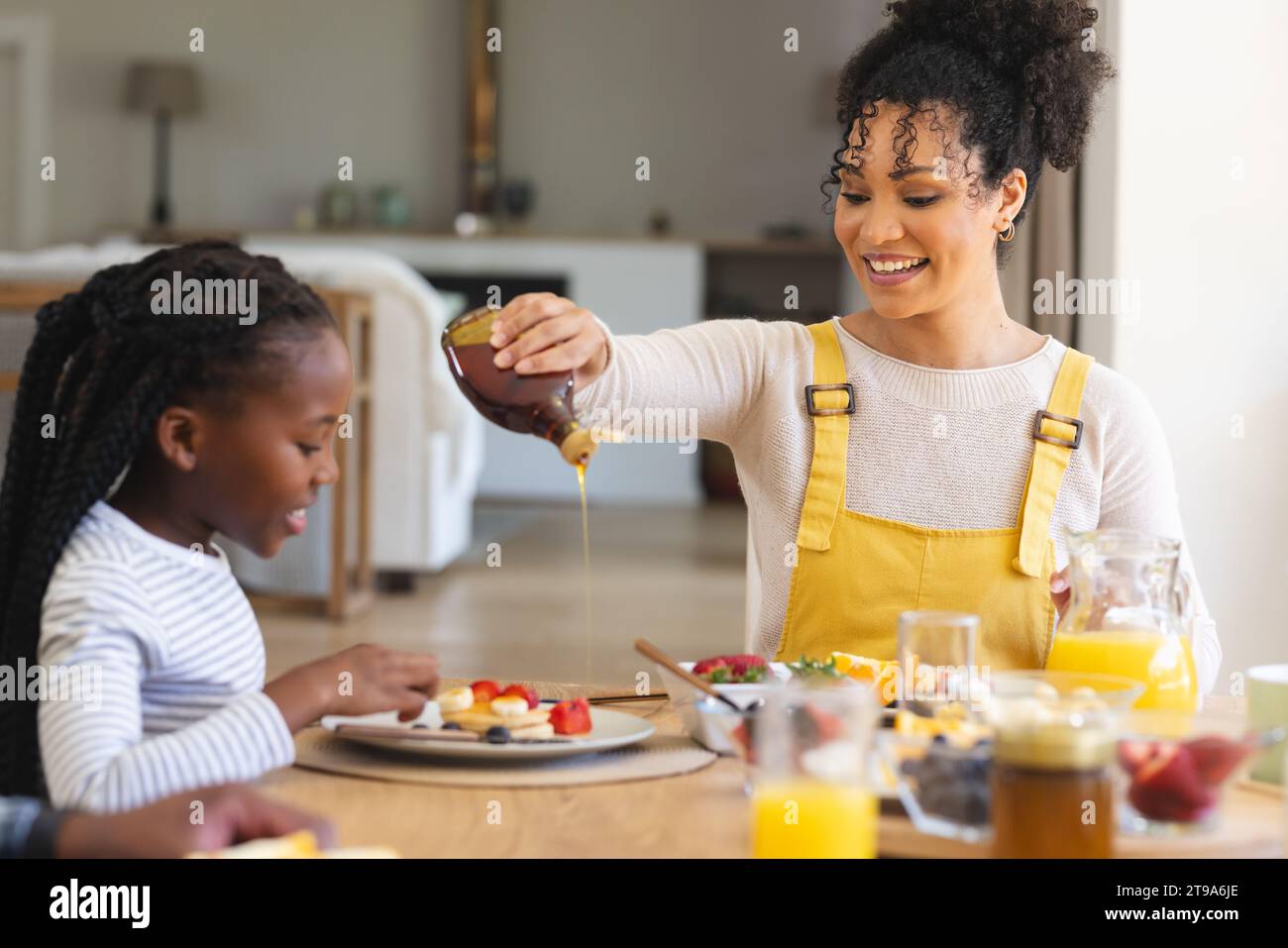 Glückliche afroamerikanische Mutter, die Ahornsirup auf Früchte gießt, im Esszimmer zu Hause, Kopierraum. Zweisamkeit, Mutterschaft, Kindheit, Essen und Trinken Stockfoto