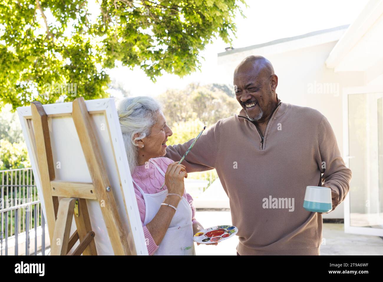 Glückliches, vielseitiges Seniorenpaar, das sich auf der sonnigen Terrasse mit Gemälden umschmeichelt und sich diese ansieht. Lebensstil, Ruhestand, Seniorenleben, Natur, Kreativität, zusammen Stockfoto