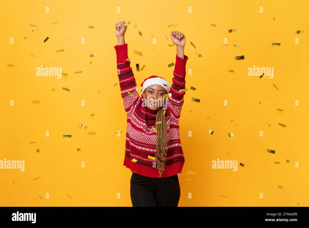 Festliches Studio fotografierte Porträt einer fröhlich lächelnden afroamerikanischen Frau, die Weihnachtspullover und Hut in gelbem Hintergrund mit Konfetti trägt Stockfoto