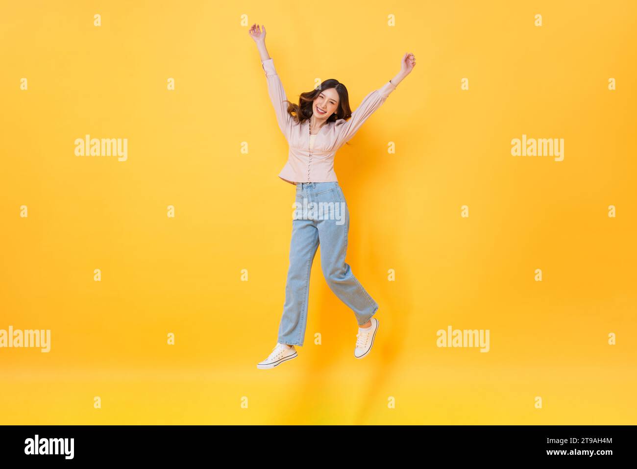 Fröhliche asiatische Frau in lässiger Kleidung lächelnd und springend mit Händen nach oben in bunter gelber Farbe isolierte Hintergrundaufnahmen im Studio Stockfoto