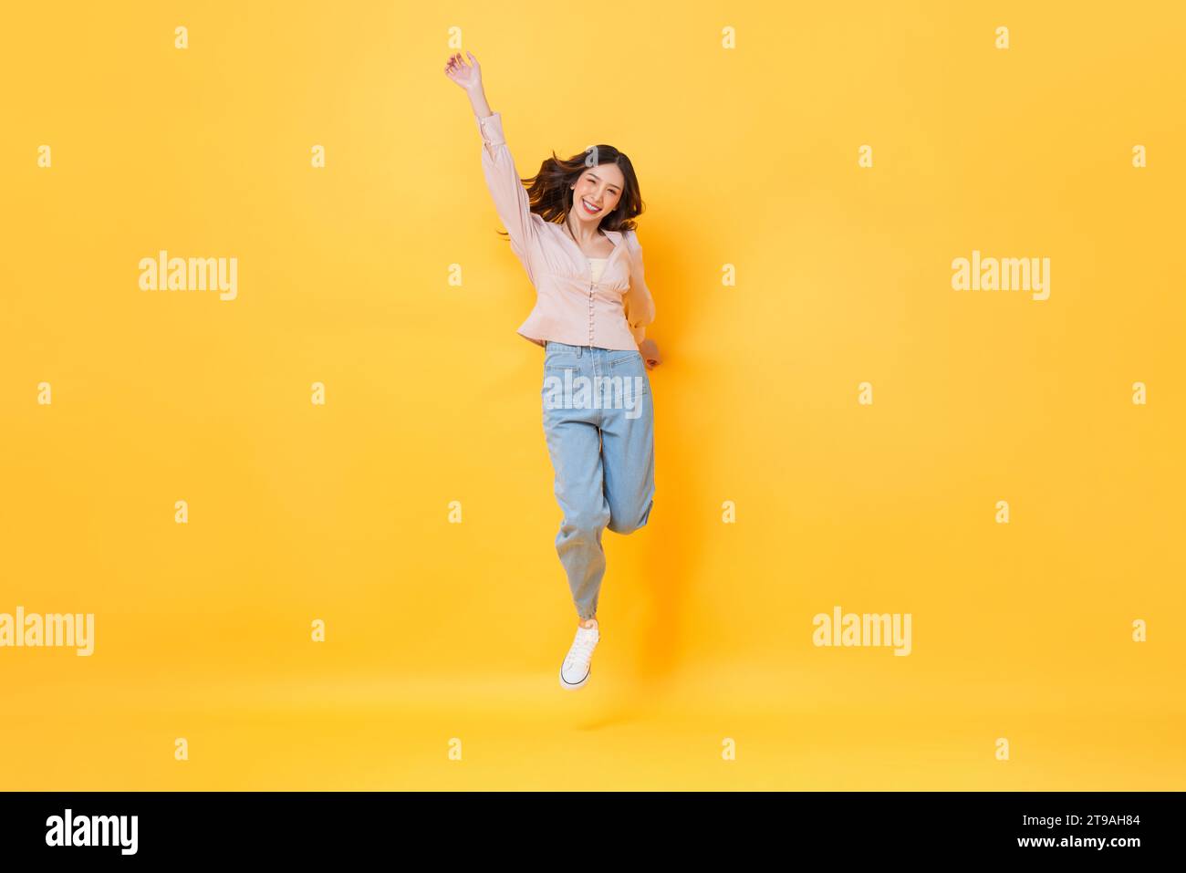 Fröhliche asiatische Frau in lässiger Kleidung lächelnd und springend mit Hand nach oben in bunter gelber Farbe isolierte Hintergrundstudio-Aufnahme Stockfoto