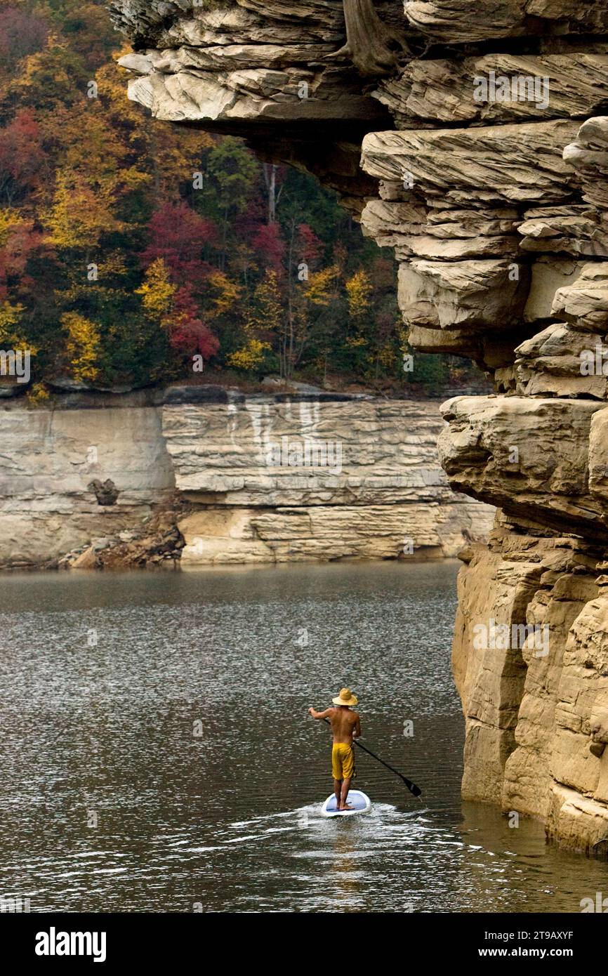 Ein Mann steht auf einem See unter großen Klippen mit Herbstfarben paddelnd auf. Stockfoto