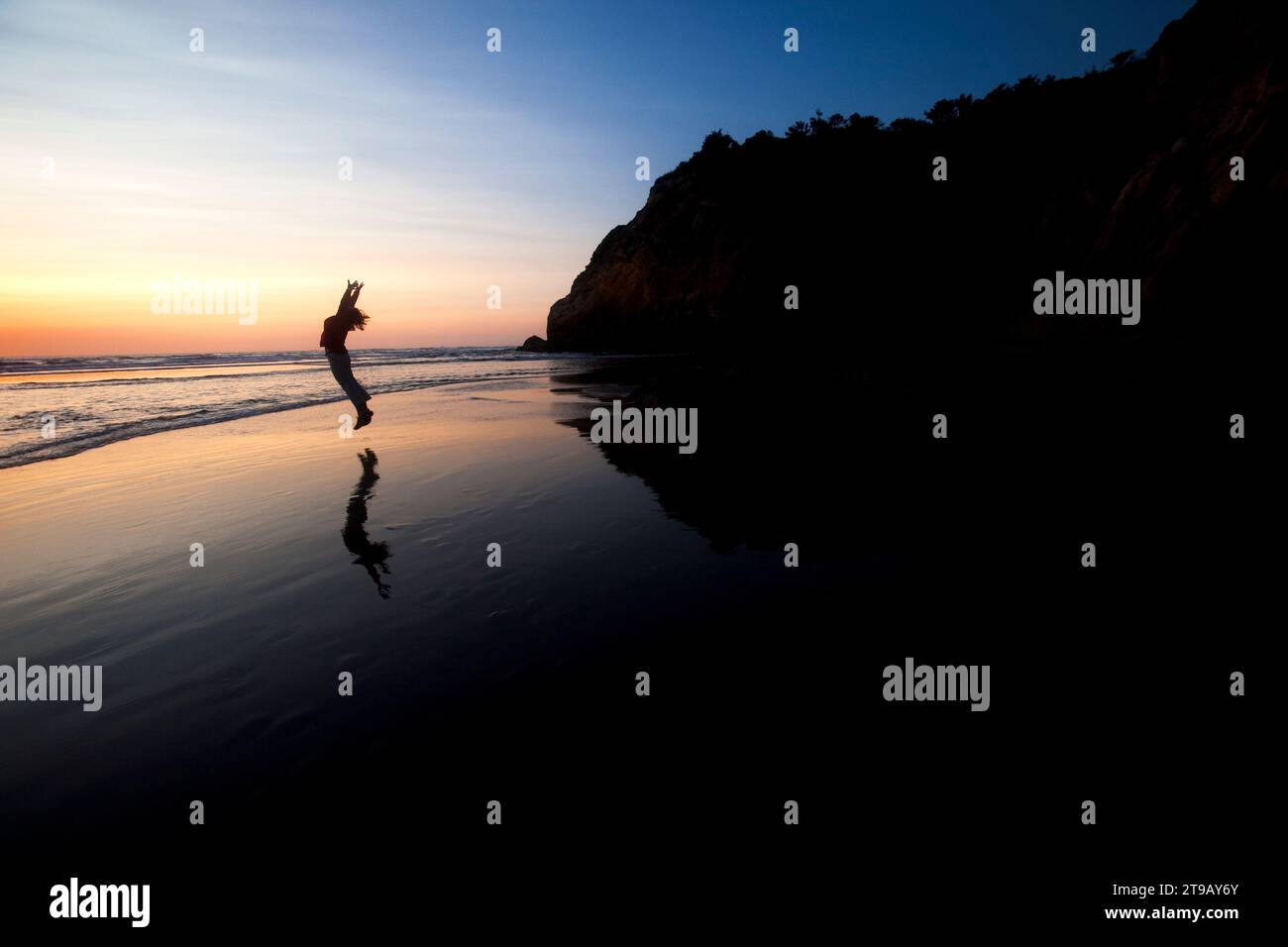 Weibliche Silhouette, die in einen Sonnenuntergang an einem Strand, umgeben von Klippen, springt. Stockfoto