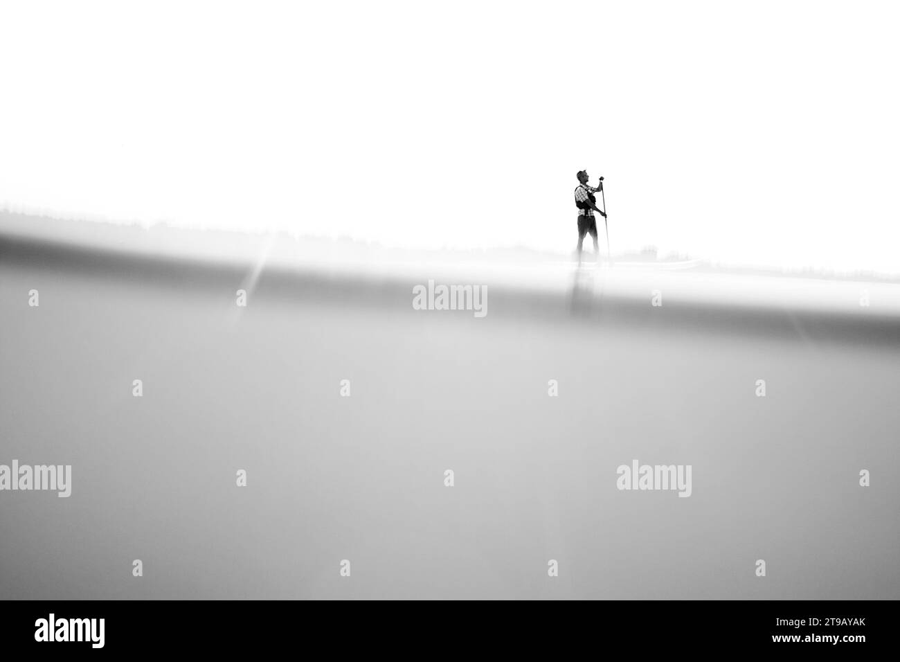 Schwarzweiß-Bild mit niedrigem Winkel und geteilter Ebene eines Mannes, der ein Stand Up Paddleboarding (SUP) gegen einen aufgeblasenen Himmel paddelt. Stockfoto
