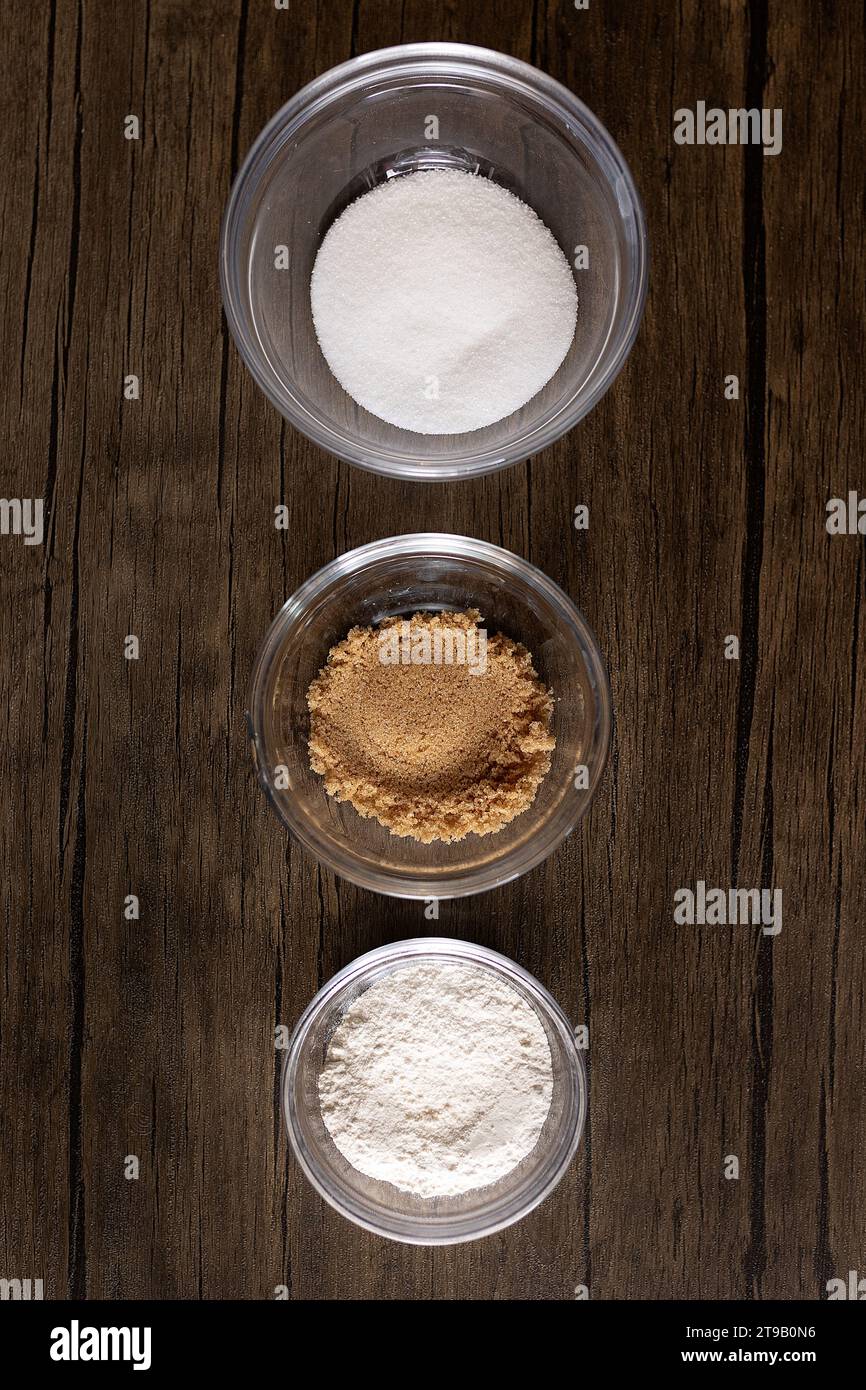Zucker, brauner Zucker und Mehl in Glasschalen gegen Holz Stockfoto