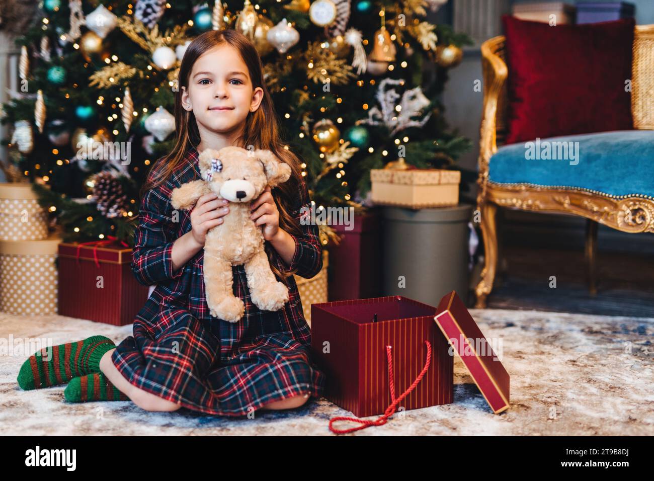 Zufriedenes Mädchen im karierten Pyjama mit einem Teddybären, einem Weihnachtsbaum und Geschenke in einem reich verzierten Zimmer Stockfoto