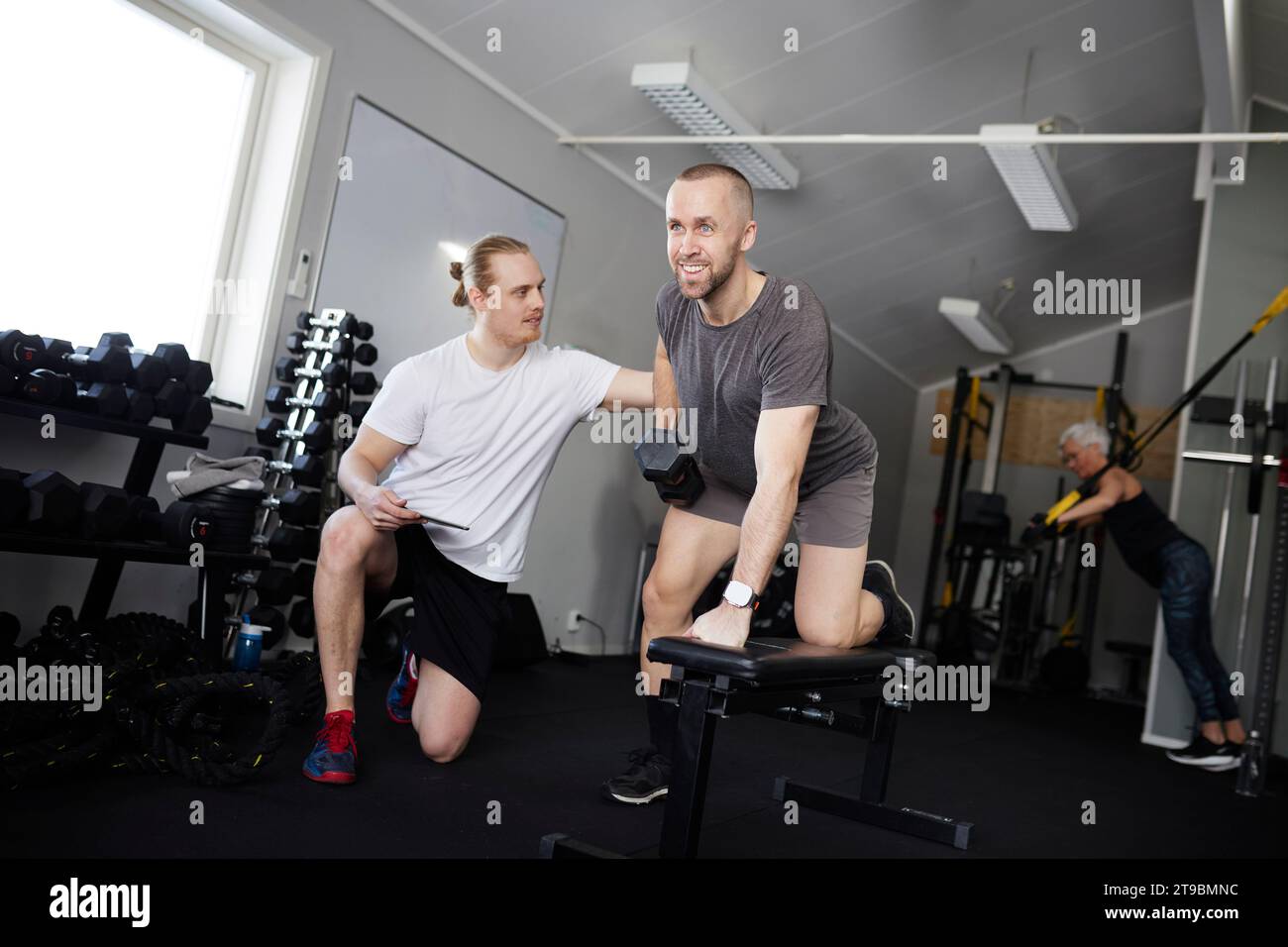 Mann, der ein Fitnessstudio ausübt, mit Trainer, der seinen Fortschritt überprüft Stockfoto