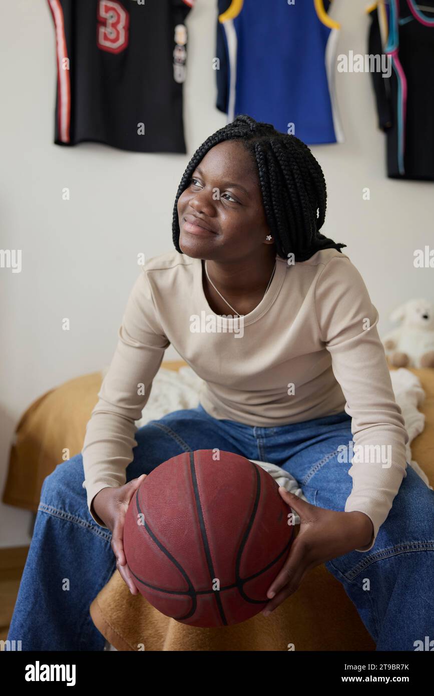 Weibliche Teenagerin, die Basketball hält, während sie im Schlafzimmer zu Hause träumt Stockfoto