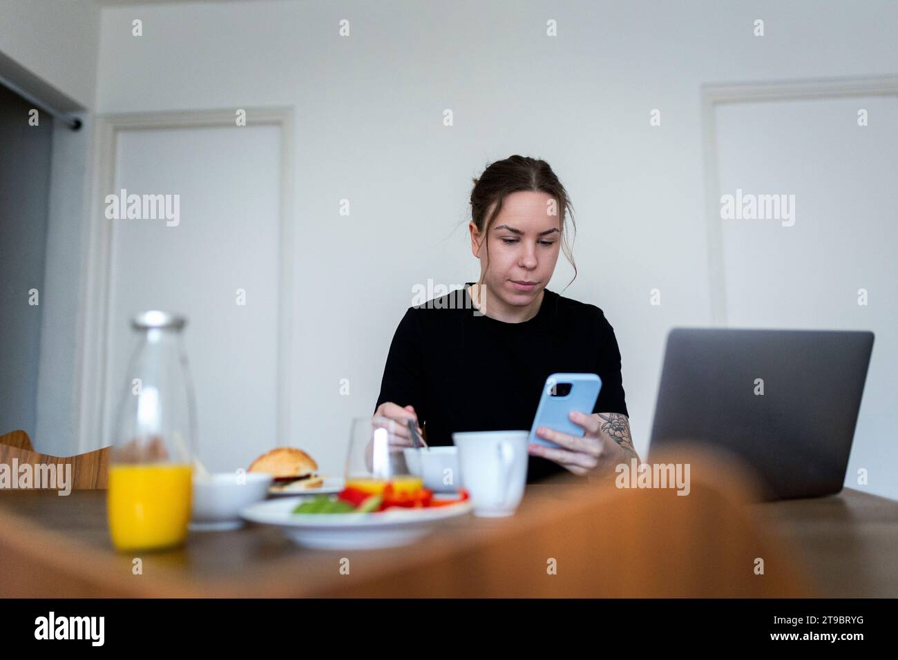 Junge Frau, die ihr Smartphone benutzt, während sie mit Laptop sitzt und am Esstisch frühstückt Stockfoto