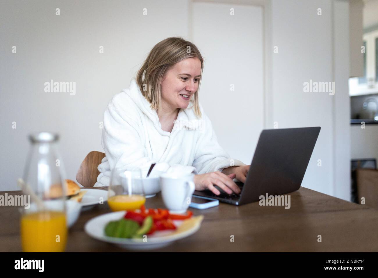 Lächelnde blonde Frau, die am Esstisch einen Laptop benutzt Stockfoto