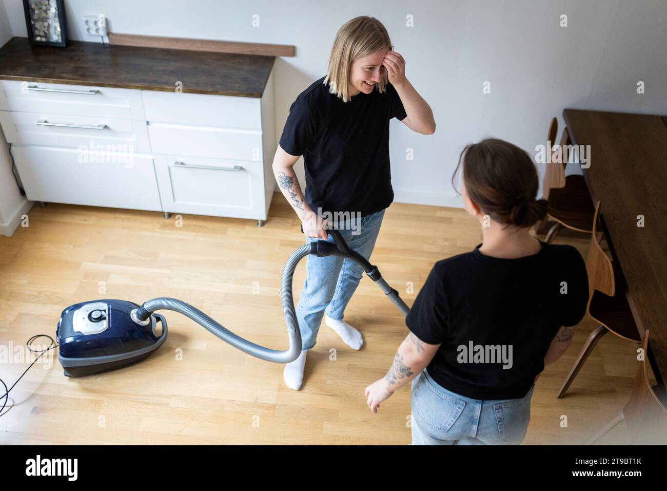 Hochwinkelansicht einer jungen lesbischen Frau, die mit ihrer Freundin spricht, während sie zu Hause mit einem Staubsauger steht Stockfoto
