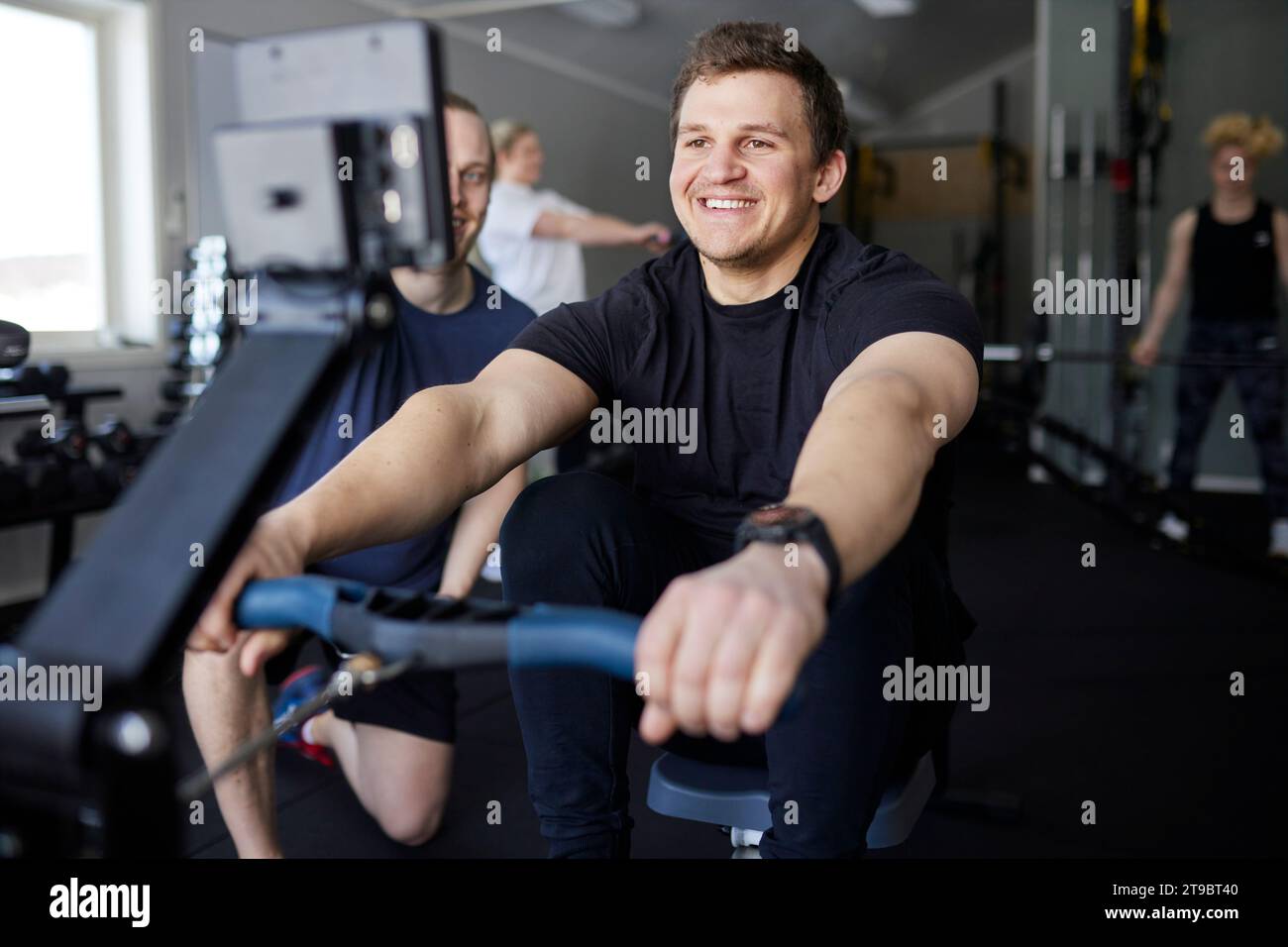 Lächelnder junger Mann, der auf Rudergerät vom Fitnesstrainer im Fitnessstudio trainiert Stockfoto