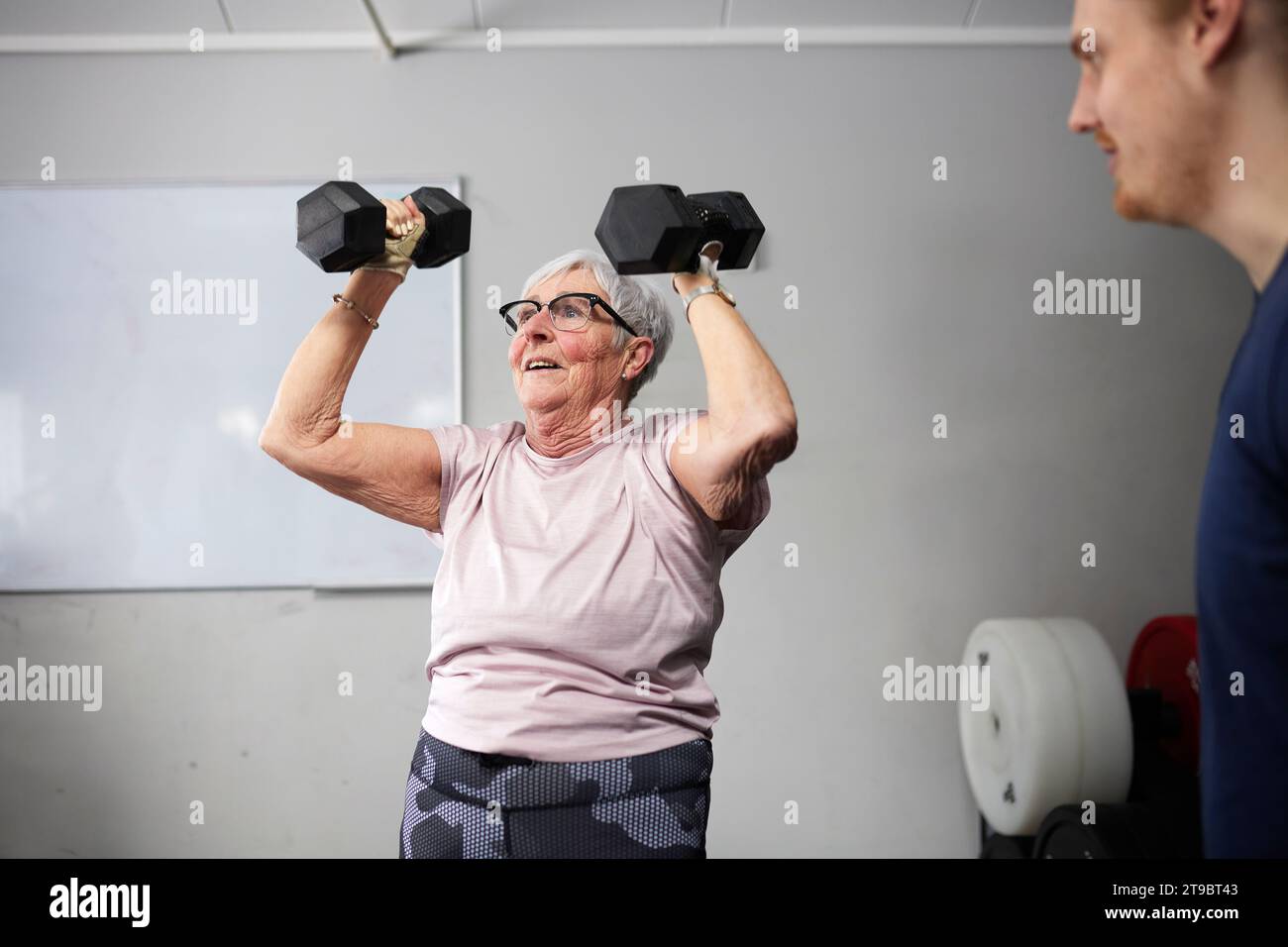 Seniorin, die mit Kurzhanteln durch männlichen Fitnesstrainer im Fitnessstudio trainiert Stockfoto