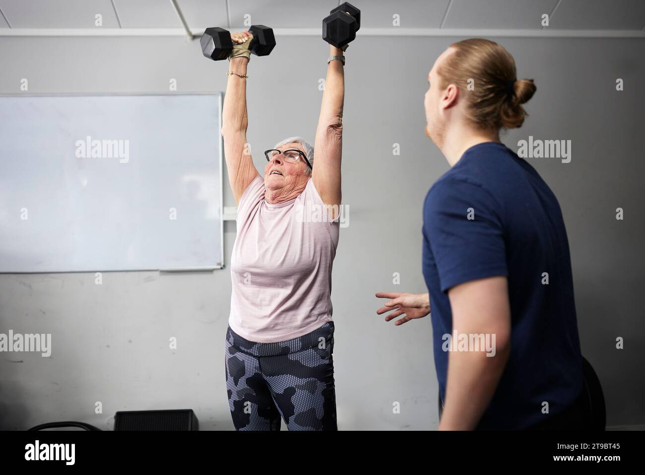 Fitnesstrainerin steht neben einer Seniorin, die im Fitnessstudio mit Kurzhanteln trainiert Stockfoto