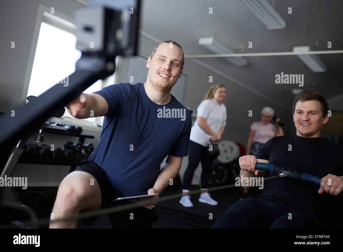 Lächelnder männlicher Fitnesstrainer mit Blick auf das Rudergerät von einem Mann, der im Health Club trainiert Stockfoto