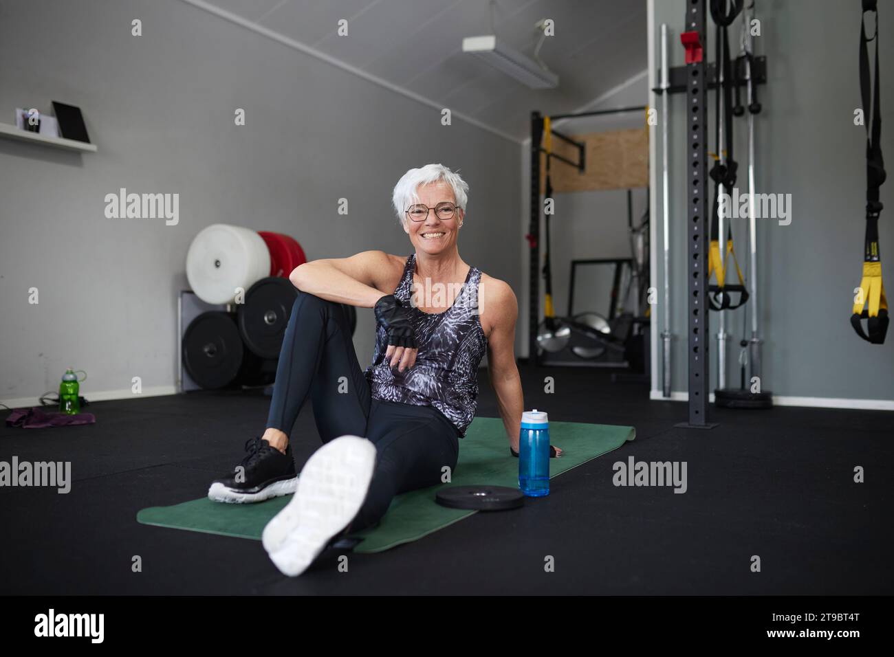 Porträt der lächelnden älteren Frau, die auf einer Trainingsmatte im Gesundheitsclub sitzt Stockfoto