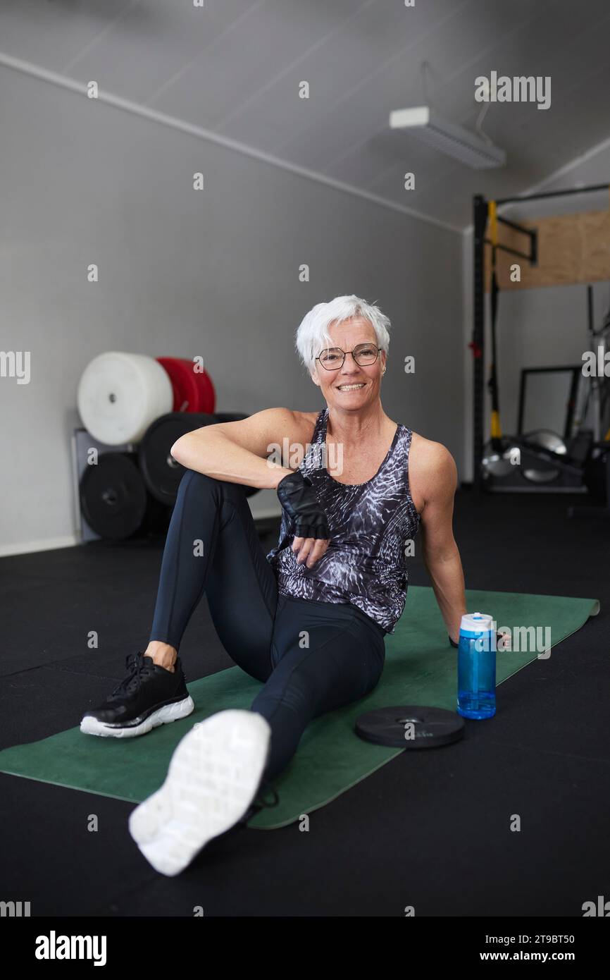 Porträt der glücklichen älteren Frau, die auf einer Trainingsmatte im Gesundheitsclub sitzt Stockfoto