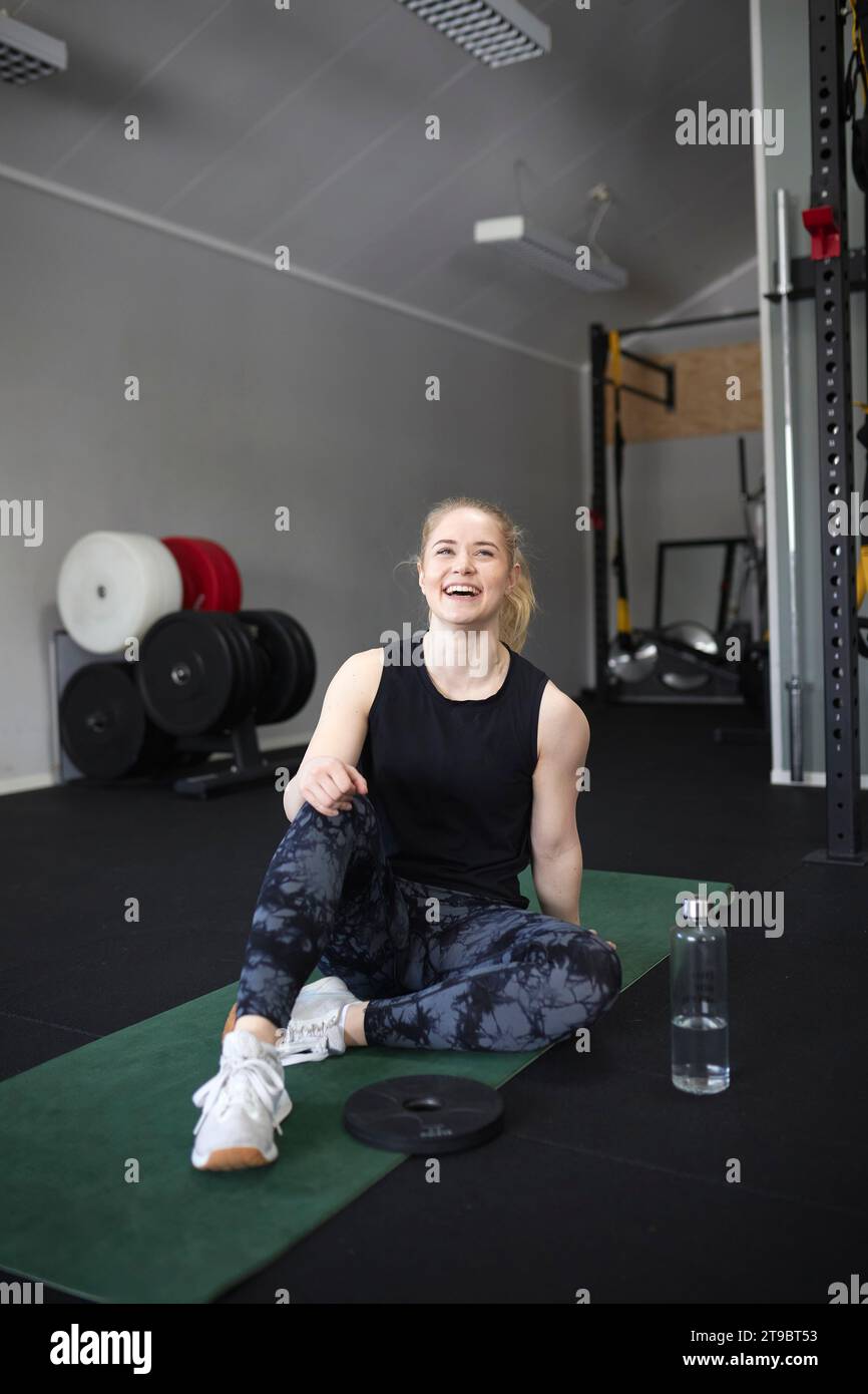Die ganze Länge einer glücklichen jungen Frau, die im Fitnessstudio auf einer Trainingsmatte sitzt Stockfoto