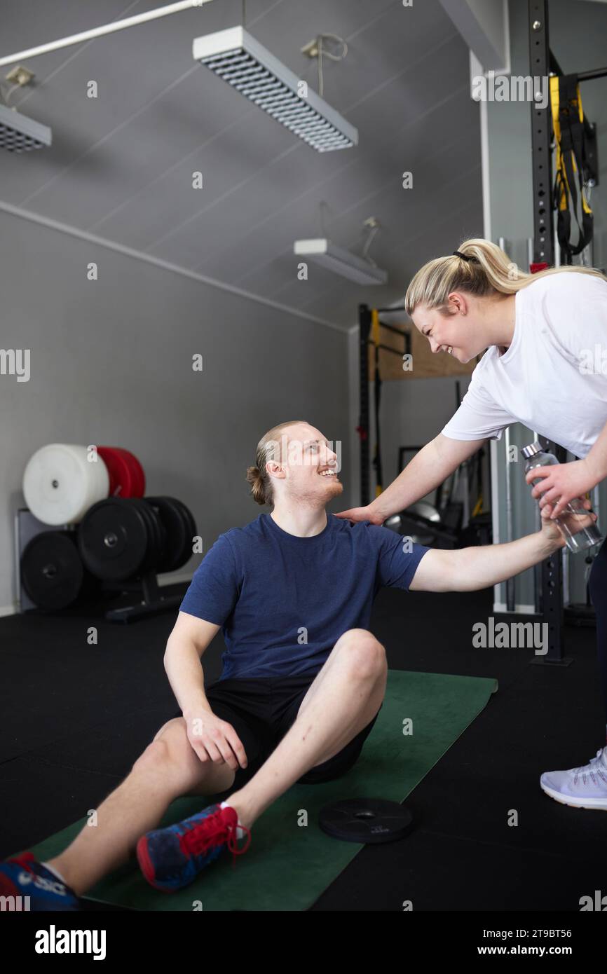 Lächelnde Frau, die dem Mann, der auf einer Trainingsmatte im Fitnessclub sitzt, eine Flasche Wasser gibt Stockfoto