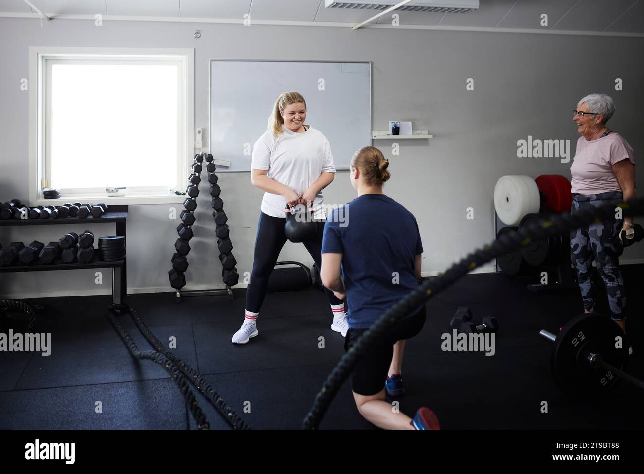 Lächelnde Frau, die mit Kettlebell vor dem Fitnesstrainer im Health Club trainiert Stockfoto