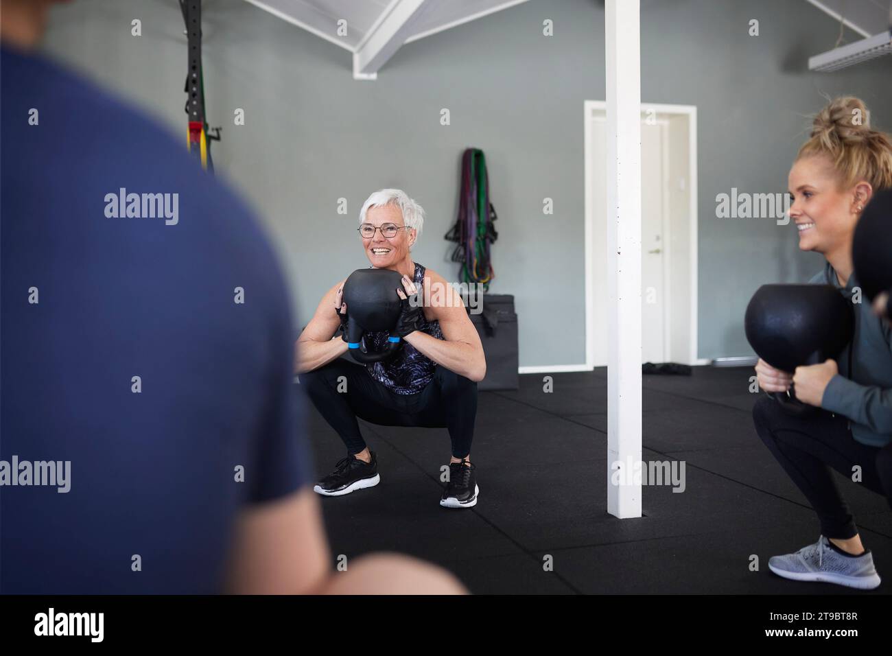 Lächelnde Frauen, die mit Kettlebells trainieren, während sie im Fitnessstudio hocken Stockfoto