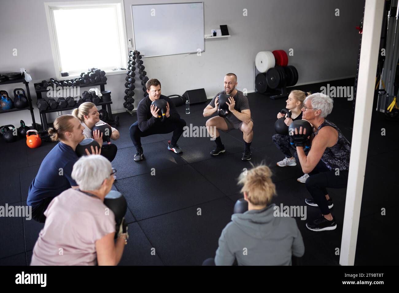Hochwinkelansicht von Personen, die mit Kettlebells trainieren, während sie im Fitnessstudio hocken Stockfoto
