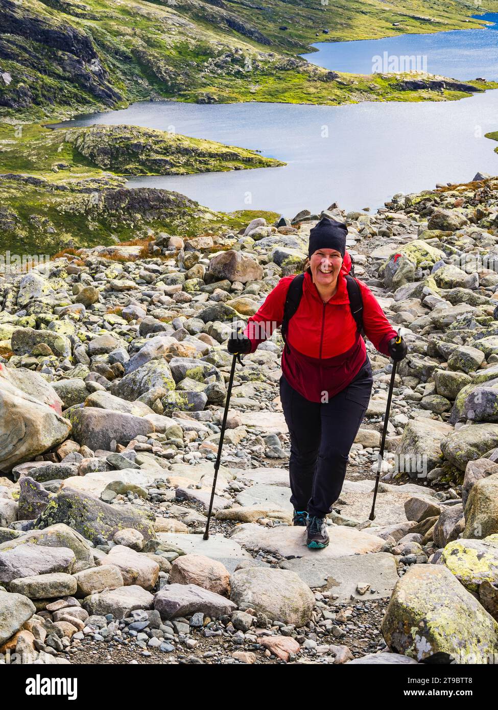 Lächelnde, reife Frau Skistöcke, die auf Felsen gegen den See laufen Stockfoto