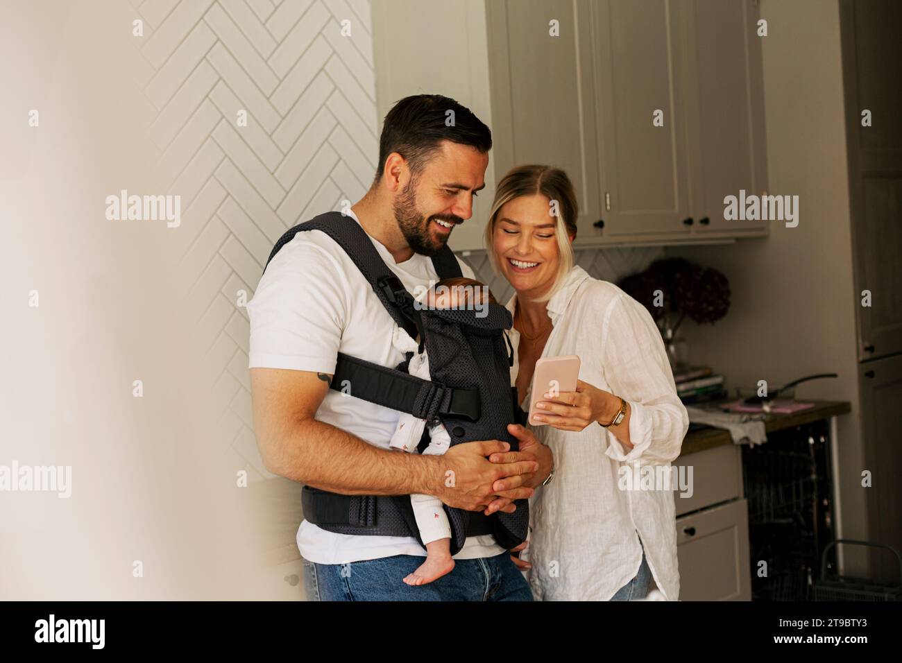 Lächelnde Frau, die ihr Smartphone mit einem Mann teilt, der zu Hause den Sohn trägt Stockfoto