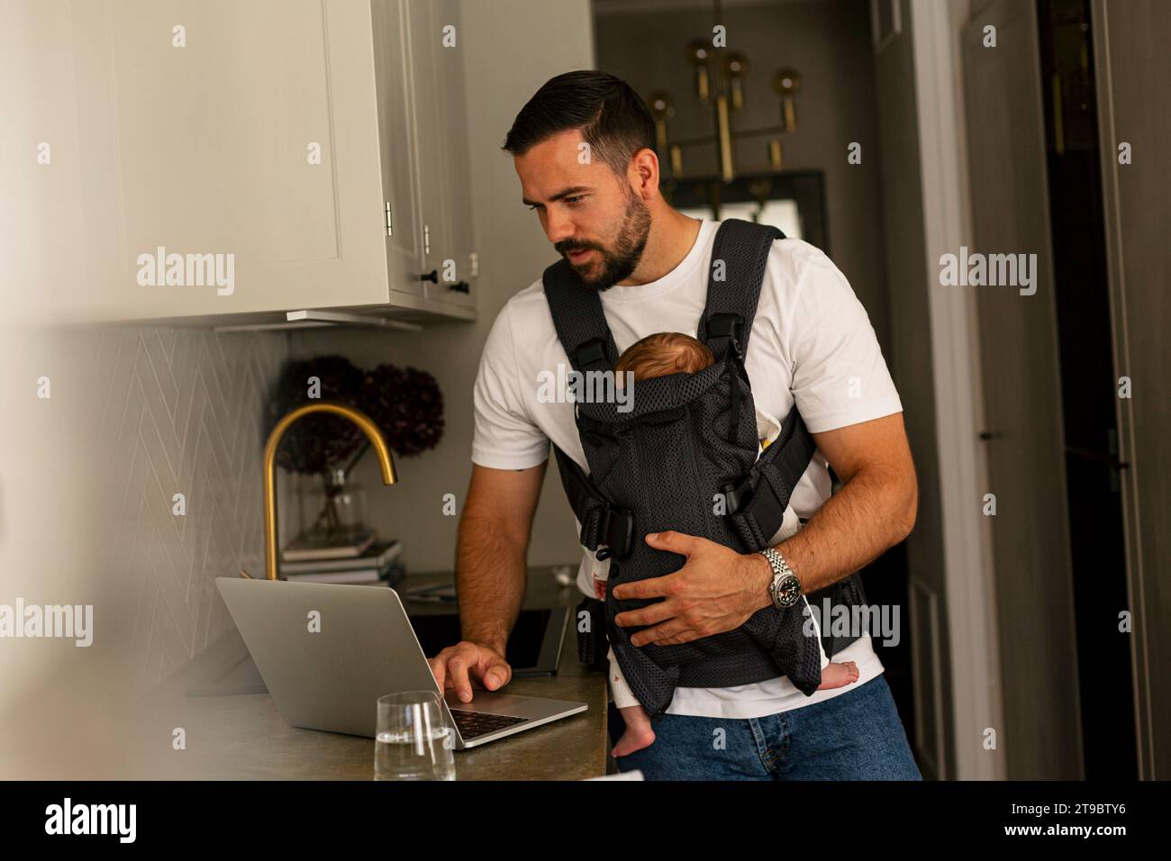 Reifer männlicher Freelancer, der mit seinem Sohn in der Trägerschaft zu Hause einen Laptop benutzt Stockfoto