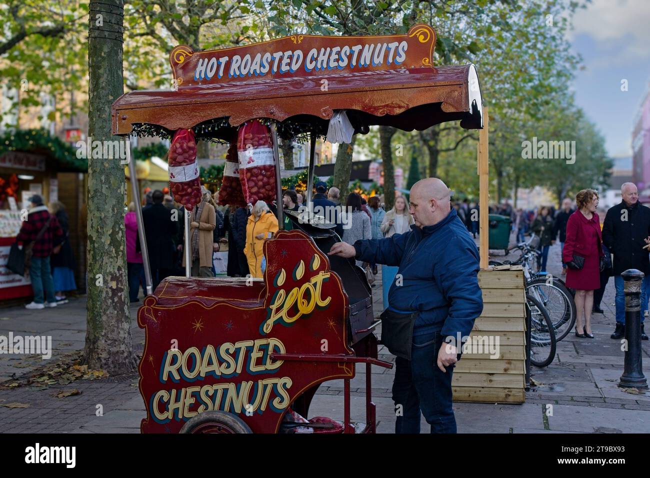 Ein festlicher rot-brauner Wagen mit einem Mann, der heisse geröstete Kastanien auf einem belebten Weihnachtsmarkt in York, Yorkshire, Großbritannien zubereitet. Stockfoto