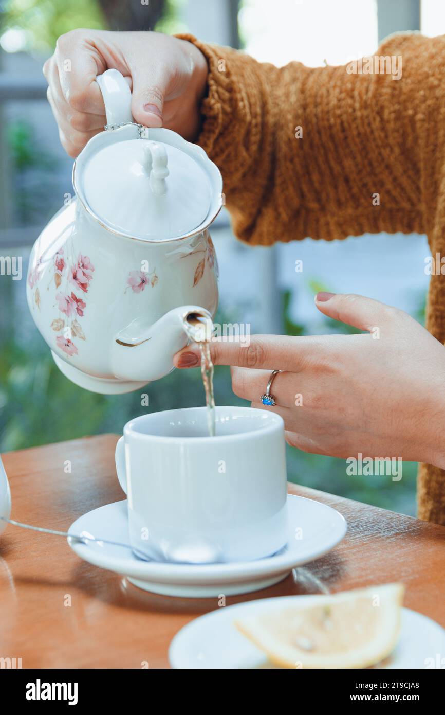 Nahaufnahme der Hände einer nicht erkennbaren kaukasischen Frau mit weißer Porzellan-Teekanne, die heissen Tee in eine weiße Porzellanschale gießt, auf Restauranttisch Stockfoto