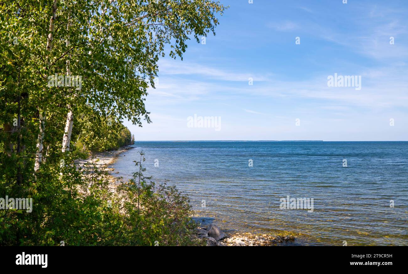 Wunderschöne ruhige See- und Seenlandschaft mit grünen Bäumen und felsigem Strand am Ufer und blauem Wasser und Himmel, mit ein paar dünnen Wolken. Stockfoto