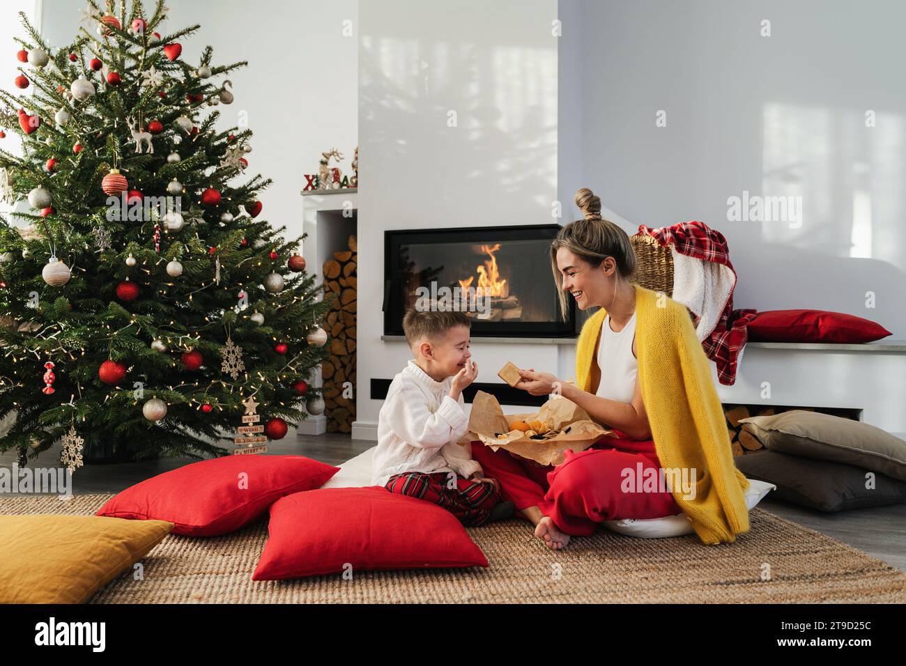 Die junge Frau und ihr kleiner Sohn sitzen neben einem glühenden Kamin in einem gemütlichen Wohnzimmer, geschmückt mit einem Weihnachtsbaum und festlichen Dekorationen, und genießen s Stockfoto