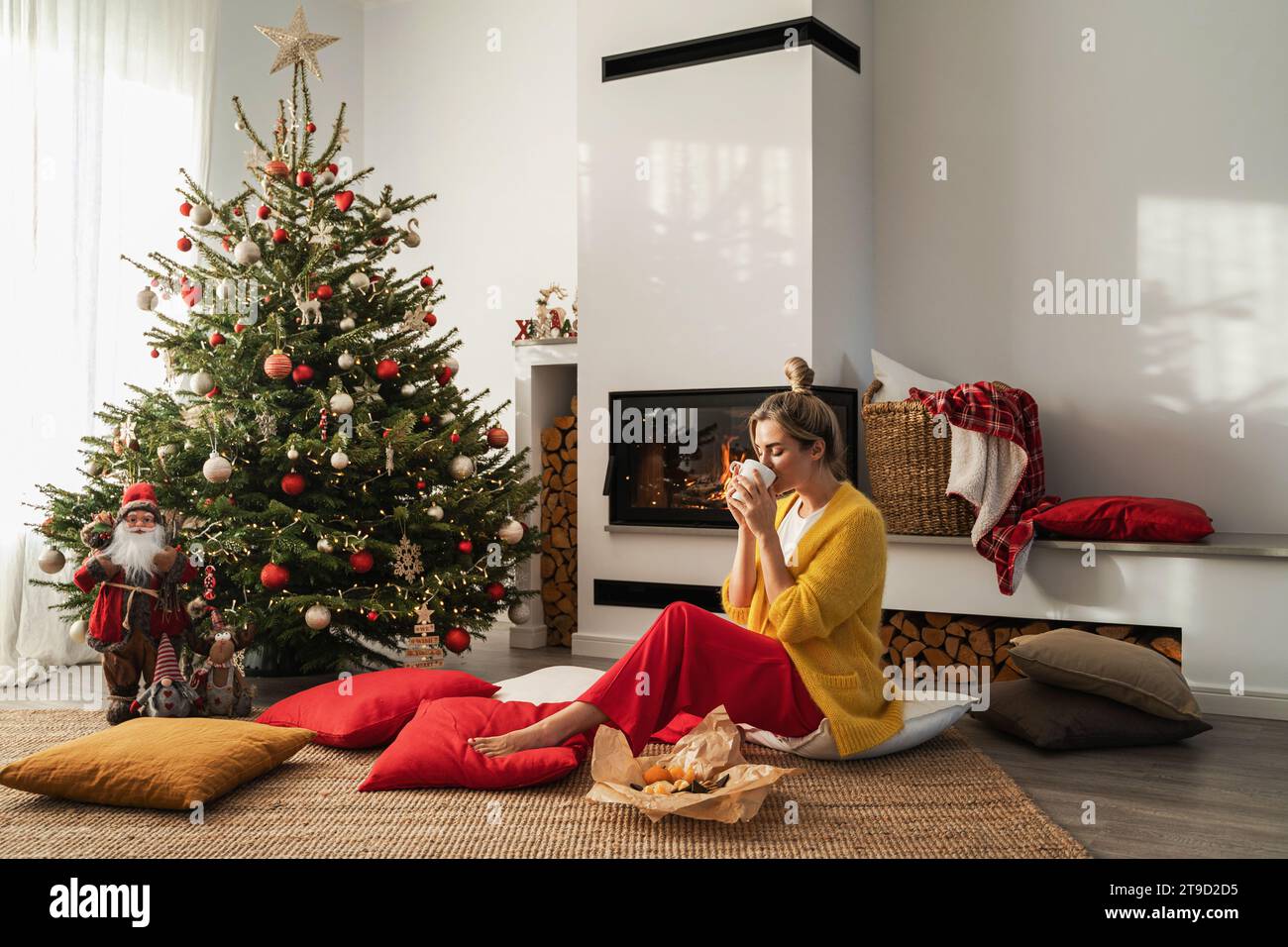 Happy Woman sitzt neben einem glühenden Kamin in einem gemütlichen Wohnzimmer, geschmückt mit einem Weihnachtsbaum und festlichen Dekorationen, und genießt eine Tasse heißes Getränk. Stockfoto