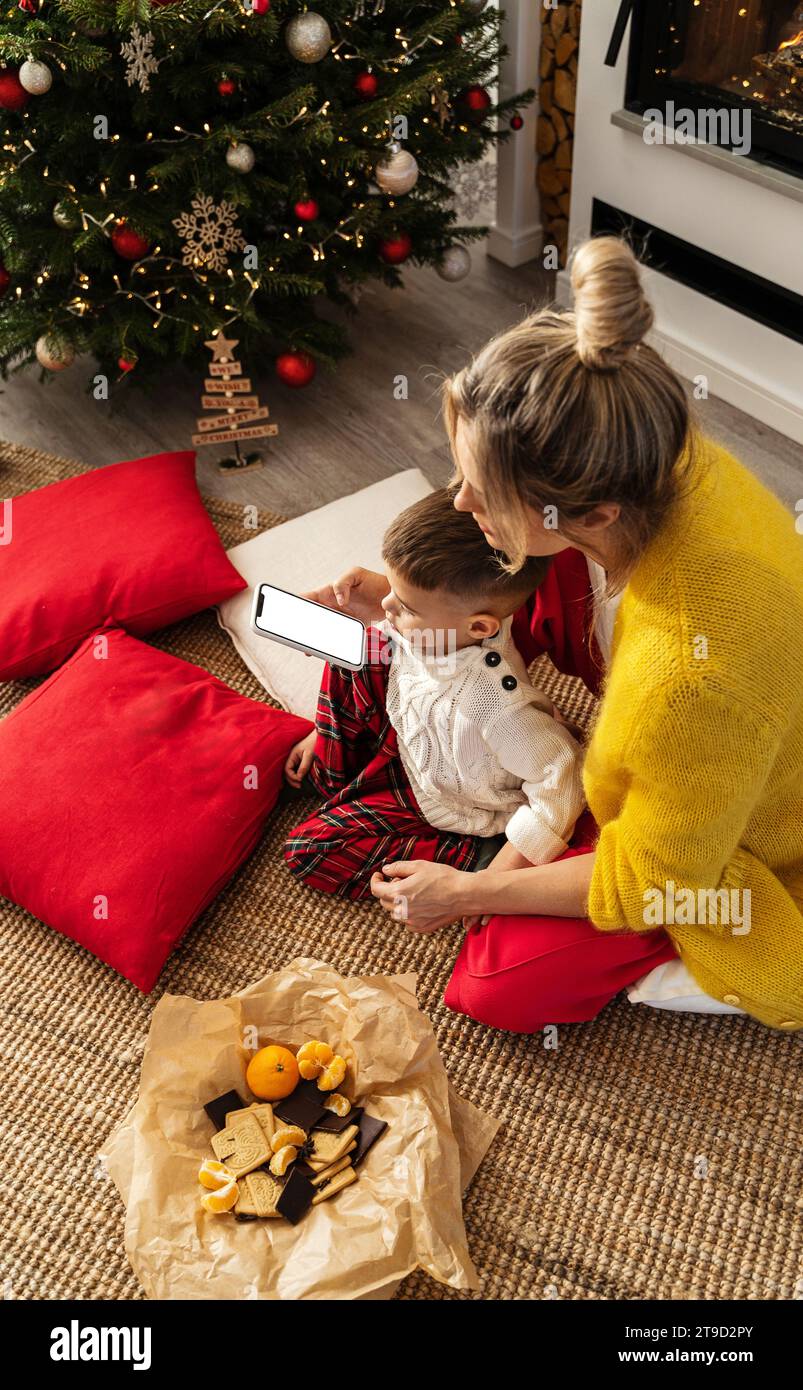 Die Frau und ihr kleiner Sohn schauen sich ein Smartphone mit einem leeren Bildschirm in einem gemütlichen Wohnzimmer an, das mit einem Weihnachtsbaum und festlichen Dekorationen geschmückt ist. Stockfoto