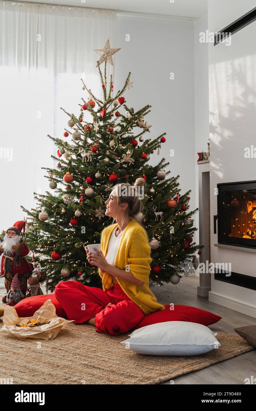 Happy Woman sitzt neben einem glühenden Kamin in einem gemütlichen Wohnzimmer, geschmückt mit einem Weihnachtsbaum und festlichen Dekorationen, und genießt eine Tasse heißes Getränk. Stockfoto