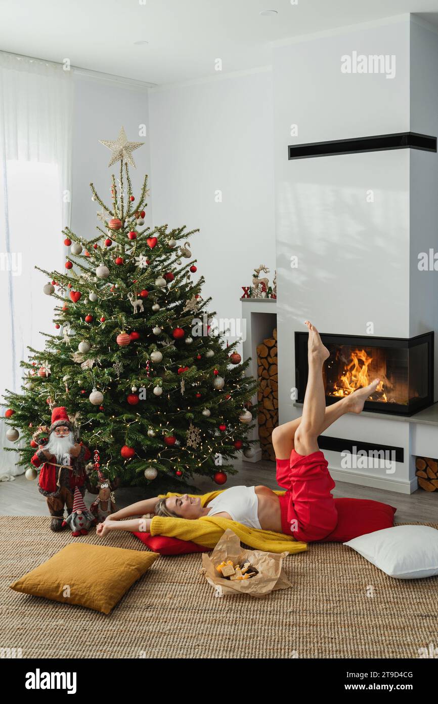 Die junge Frau liegt neben einem glühenden Kamin in einem gemütlichen Wohnzimmer, geschmückt mit einem Weihnachtsbaum und festlichen Dekorationen. Stockfoto