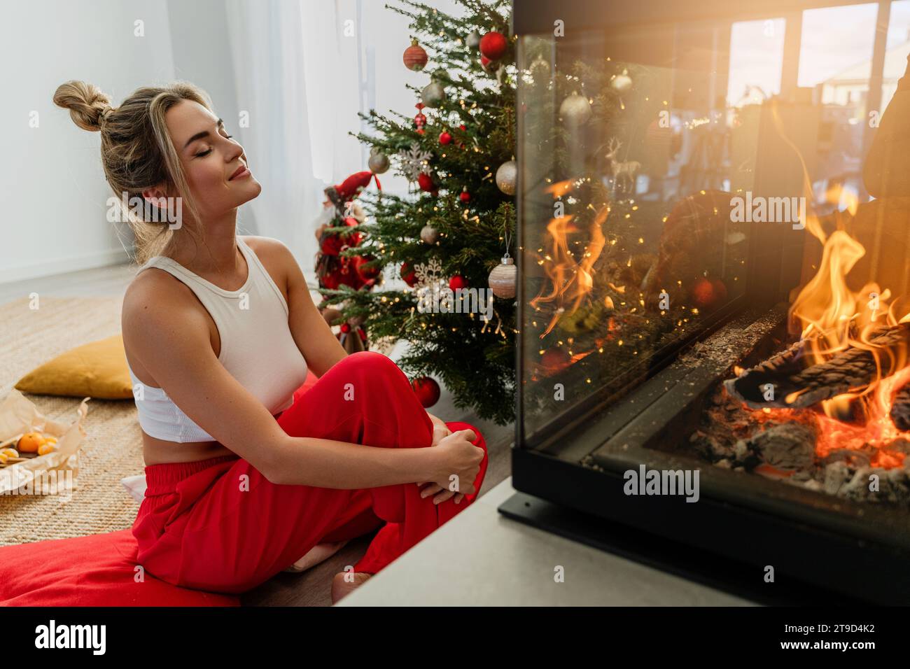 Die junge Frau sitzt neben einem glühenden Kamin in einem gemütlichen Wohnzimmer, das mit einem Weihnachtsbaum und festlichen Dekorationen geschmückt ist. Stockfoto