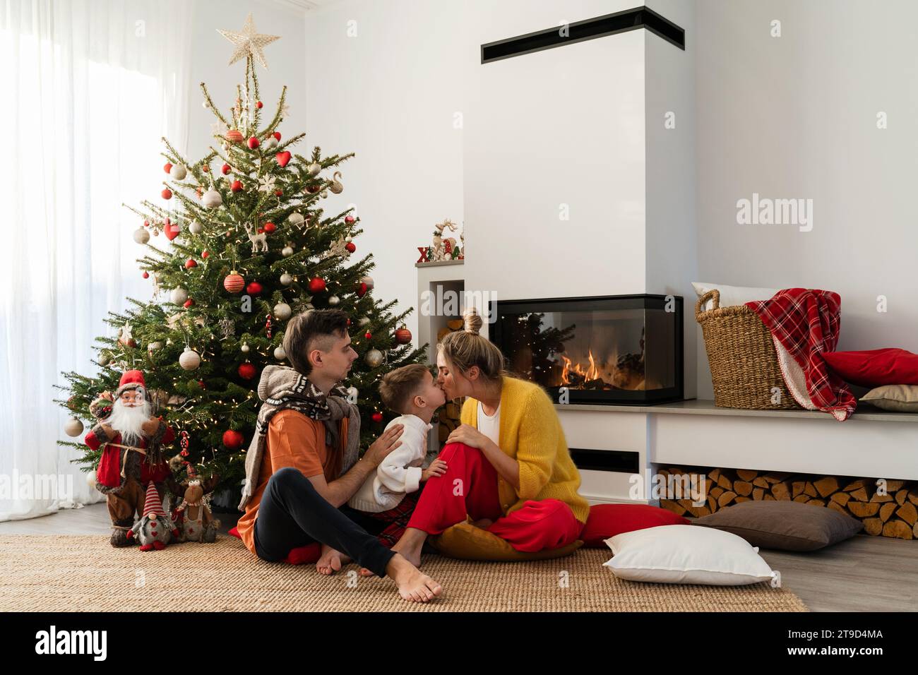 Junge glückliche Familie sitzen neben einem glühenden Kamin in einem gemütlichen Wohnzimmer, geschmückt mit einem Weihnachtsbaum und festlichen Dekorationen. Stockfoto